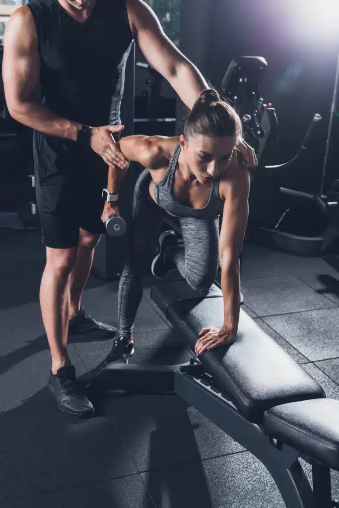 Coach guidant une femme dans l'exécution d'un exercice de musculation avec haltères lors d'une séance de coaching personnalisé.